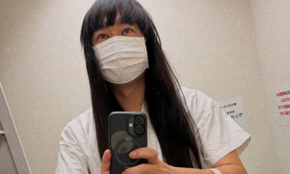 ネコニスズ・舘野忠臣(赤ちゃん)の病院での自撮り写真。ロング黒髪に前髪、白いマスクと医療用ガウン姿でスマートフォンを持ち、点滴を付けた状態。内視鏡検査後の様子を捉えたインスタグラム投稿より