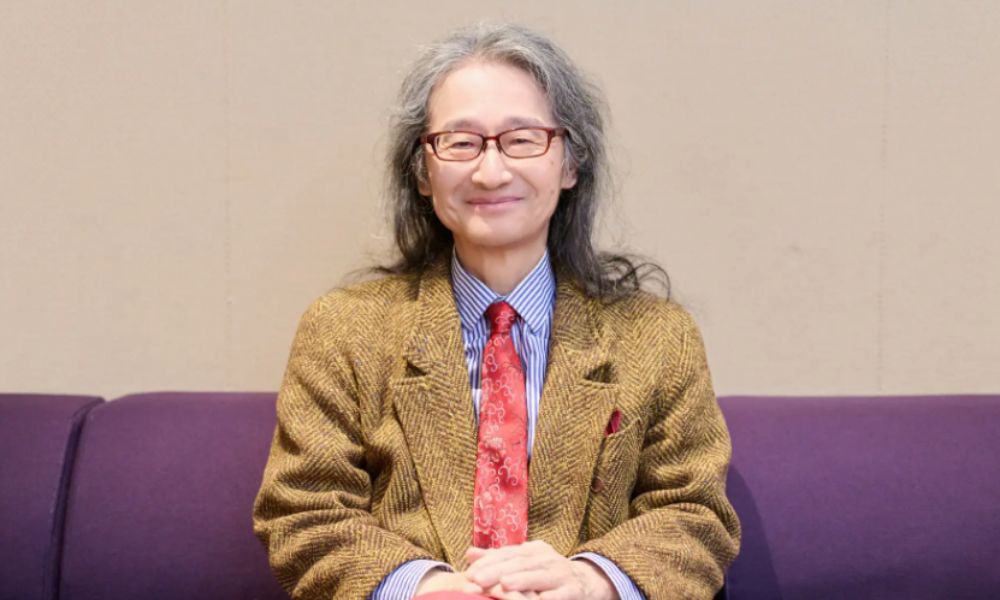 作曲家・鷺巣詩郎のポートレート。長髪に眼鏡、茶色のジャケットと赤いネクタイ姿。『エヴァンゲリオン』劇伴で知られ、父はピー・プロダクション創業者うしおそうじ