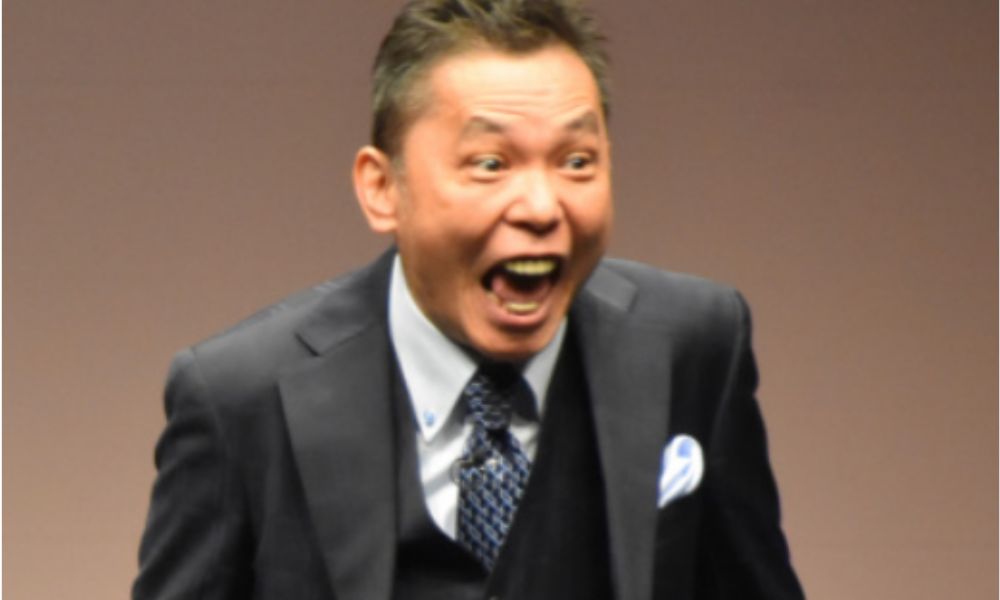爆笑問題・太田光がスーツ姿で口を大きく開けた激しい表情の写真。ダークスーツに青系の柄物ネクタイとポケットチーフを合わせ、ステージ上で感情を爆発させている様子。オリコンニュースより