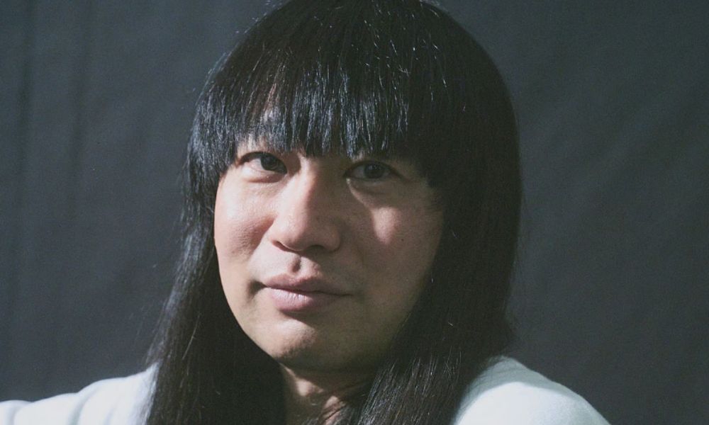 ネコニスズ・舘野忠臣(赤ちゃん)のロング黒髪ポートレート写真。前髪を切り揃えたストレートの黒髪ロングヘアが特徴の42歳お笑い芸人。白いシャツ姿でやや微笑む表情。2026年お笑いナタリー記事より